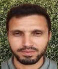 Homme avec barbe, vêtu d'une chemise grise, regard sérieux, fond vert.